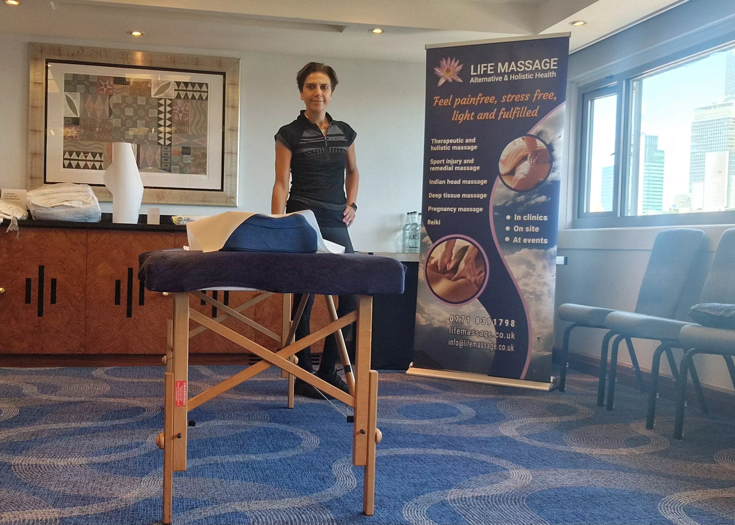 On site office setting 20240912 On-site treatments in companies. Life Massage therapist, standing beside a massage couch, set up in company office on site.